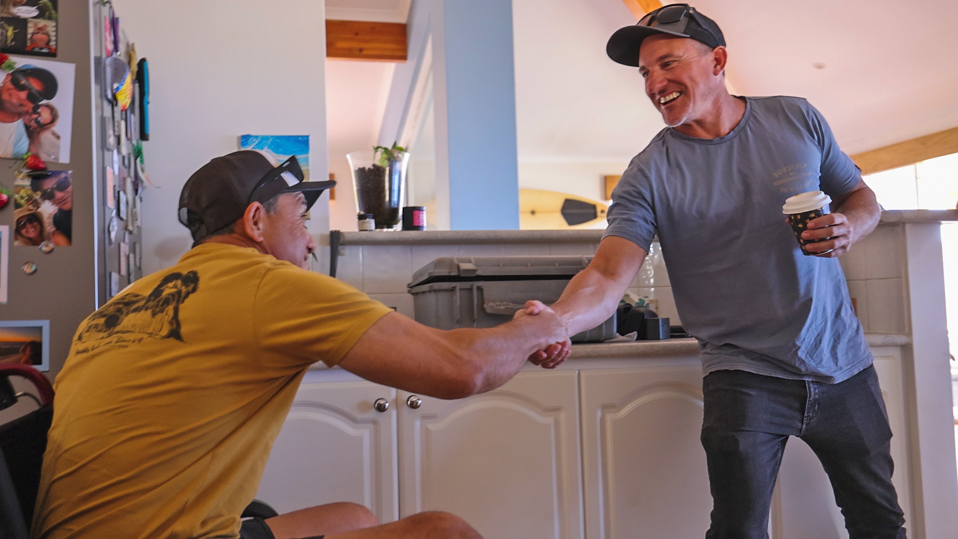 A man in a wheel chair and a man standing shake hands, smiling, in a kitchen.
