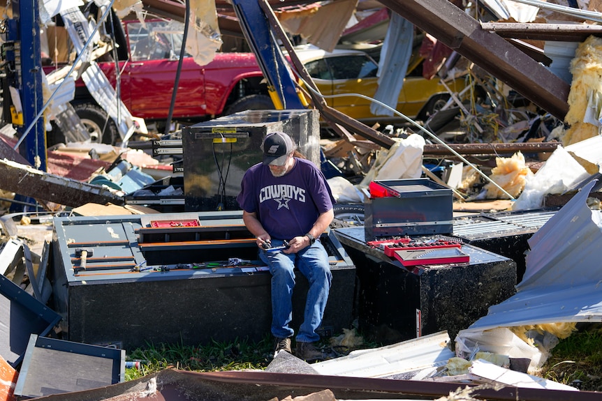 A man sits in the middle of storm damage and looks at the devastation. 