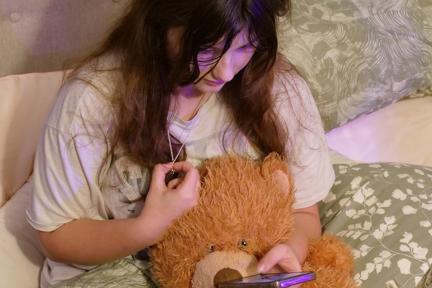 Young girl cuddles a teddy bear on phone in bed