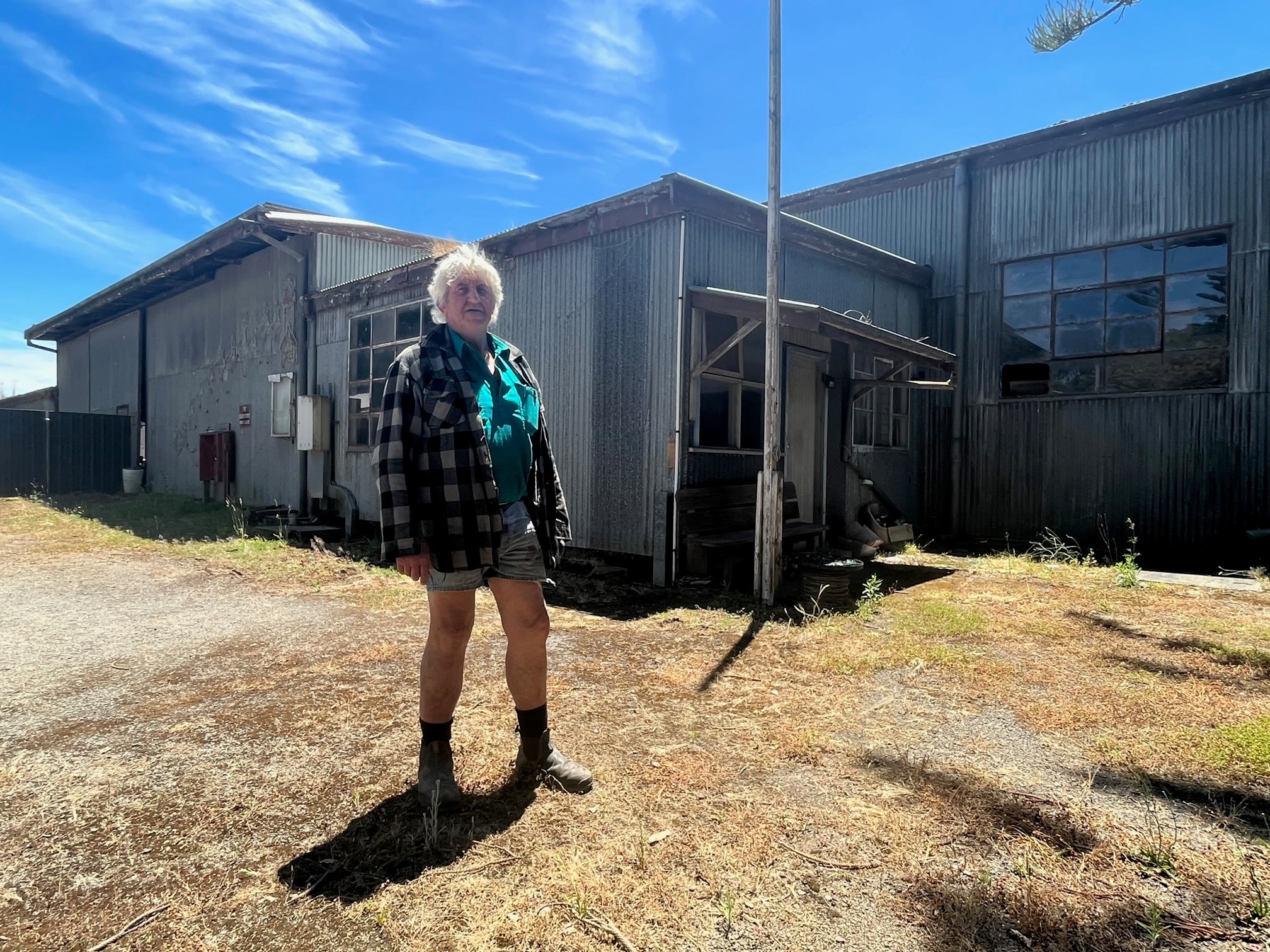 A man with white hair wearing a flannel shirt and shorts in front of a corrugated iron building