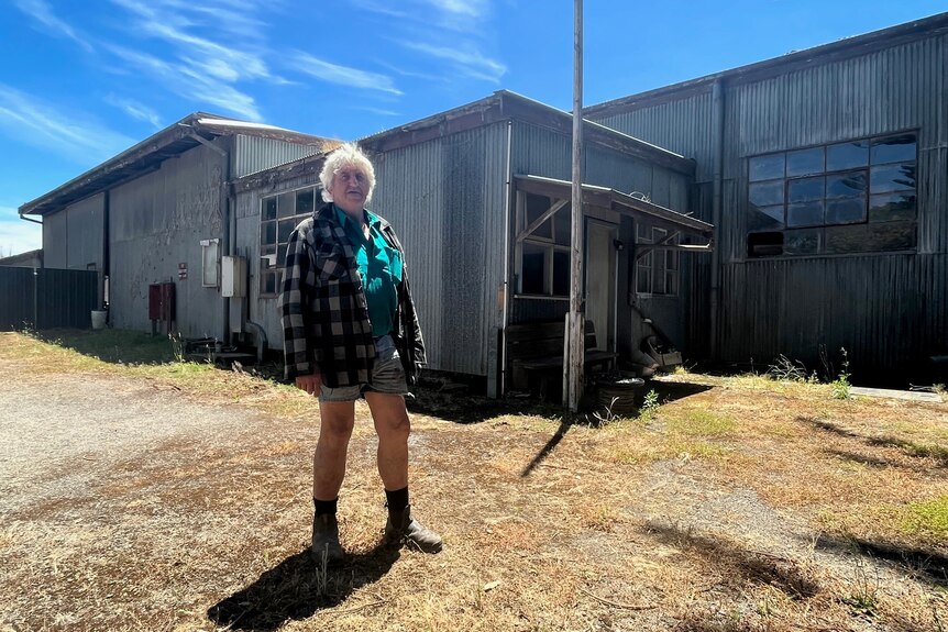 A man with white hair wearing a flannel shirt and shorts in front of a corrugated iron building