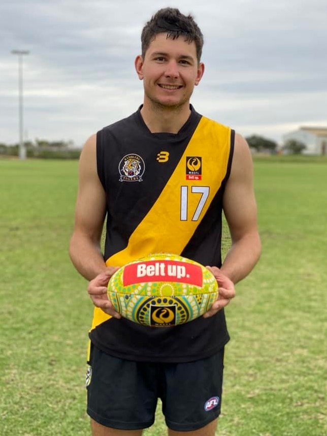 A man in footy gear holding a football smiles at the camera.