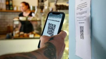 A man scans a QR code with his phone. Behind him, there is a cafe worker at a counter.