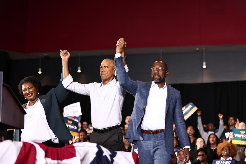 Obama holds hands and raises them in the air with Raphael Warnock and Stacey Abrams. 