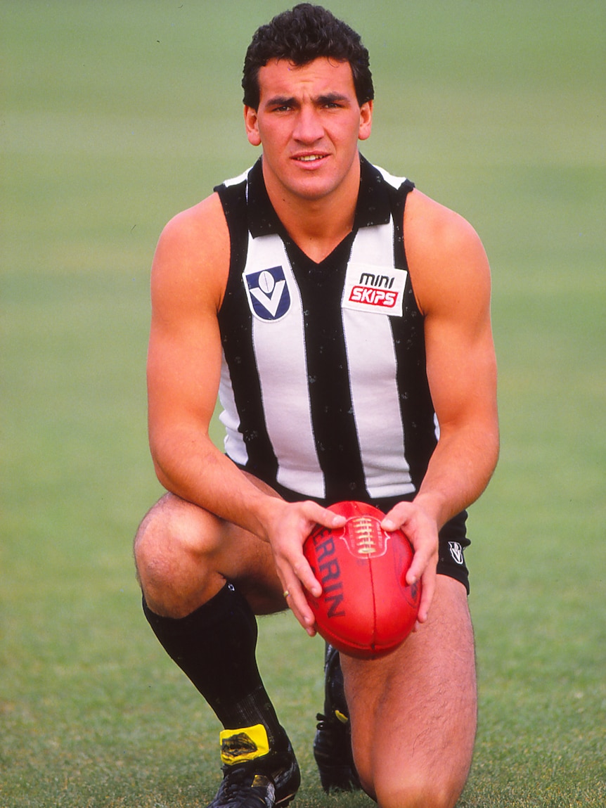 A Collingwood VFL player poses for a photograph holding a red football while wearing Collingwood's playing strip.