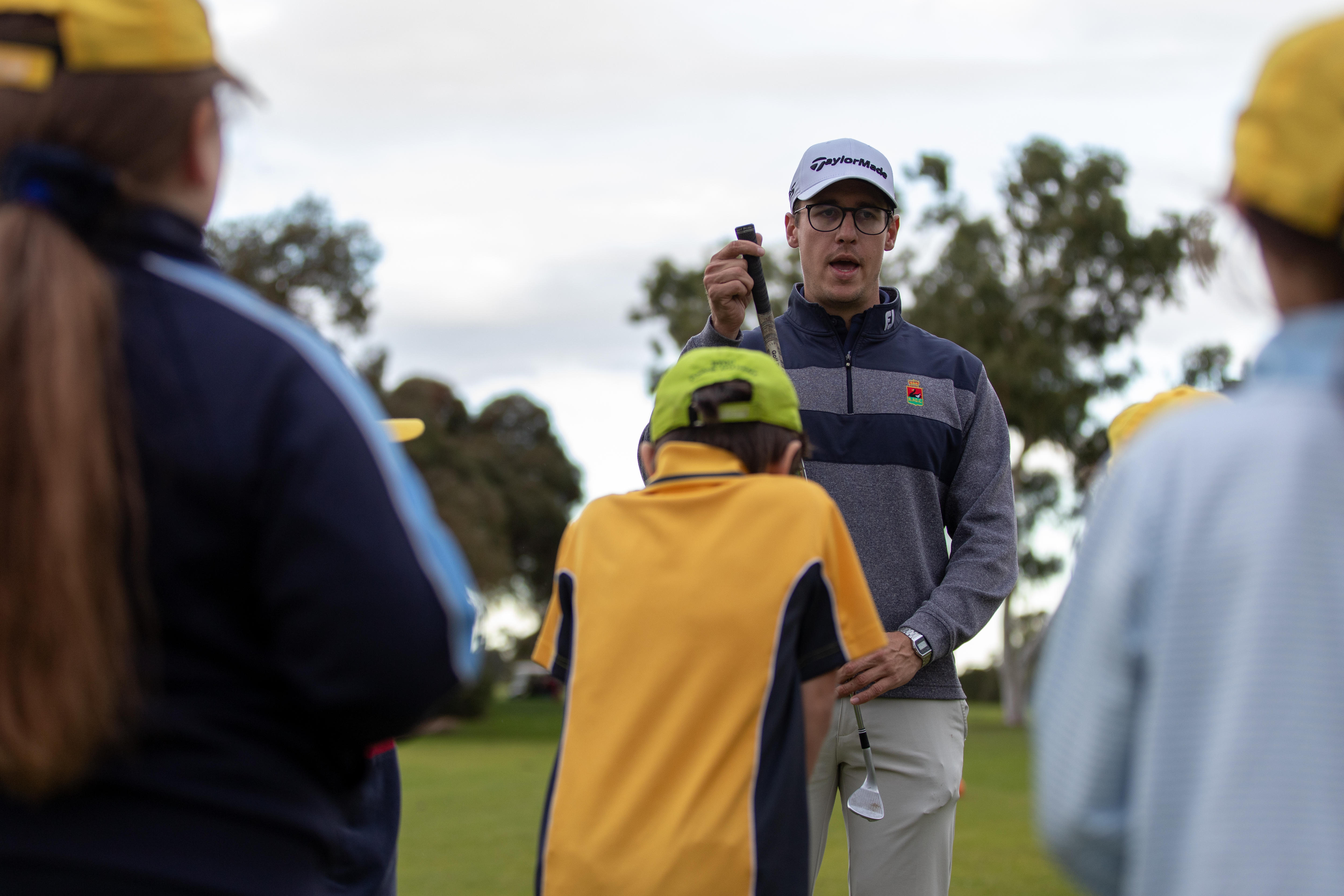 Man holds golf club while speaking in front of group of children.