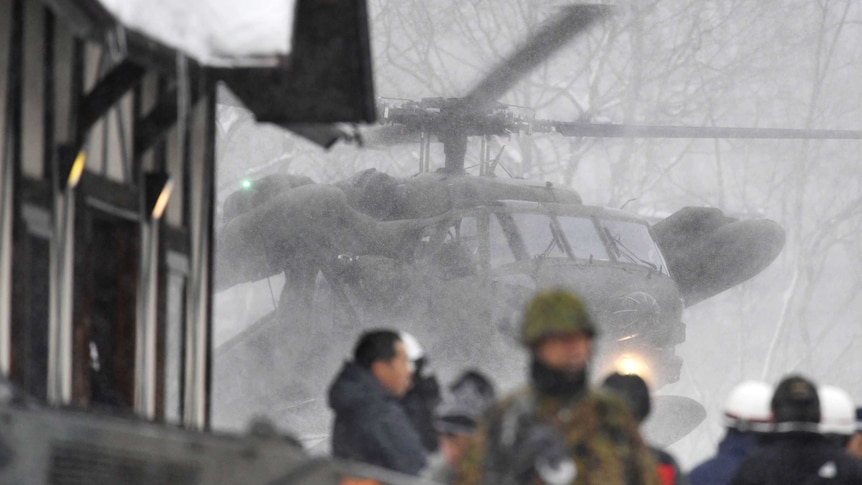 A military helicopter hovers in the background in snowy conditions as emergency responders work in the foreground
