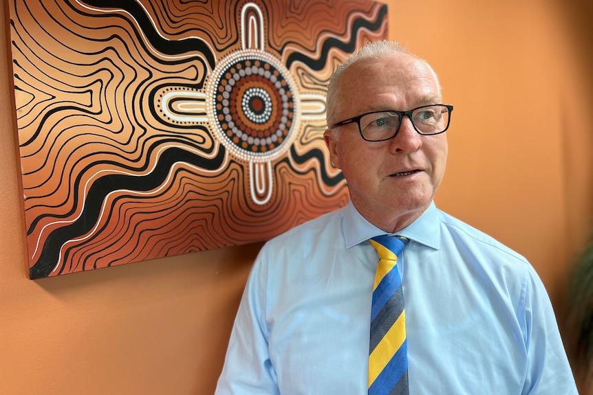A grey-haired man standing in front of an Indigenous artwork in an office.