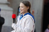 Greta Thunberg is holding a microphone as she pulls a folded piece of paper out of her pocket