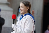 Greta Thunberg is holding a microphone as she pulls a folded piece of paper out of her pocket