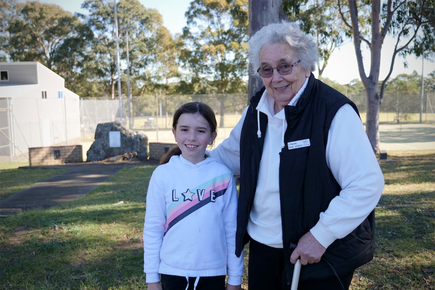 A young girl with brown hair smiles beside an elderly woman with glasses and a walking stick