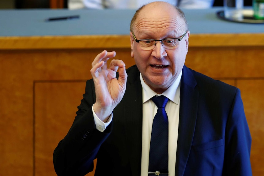 A bald man in a suit and tie makes a potentially inflammatory hand gesture.