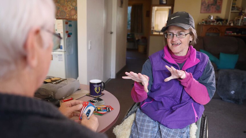 A middle-aged woman with hydrocephalus plays UNO with an older man around a table.