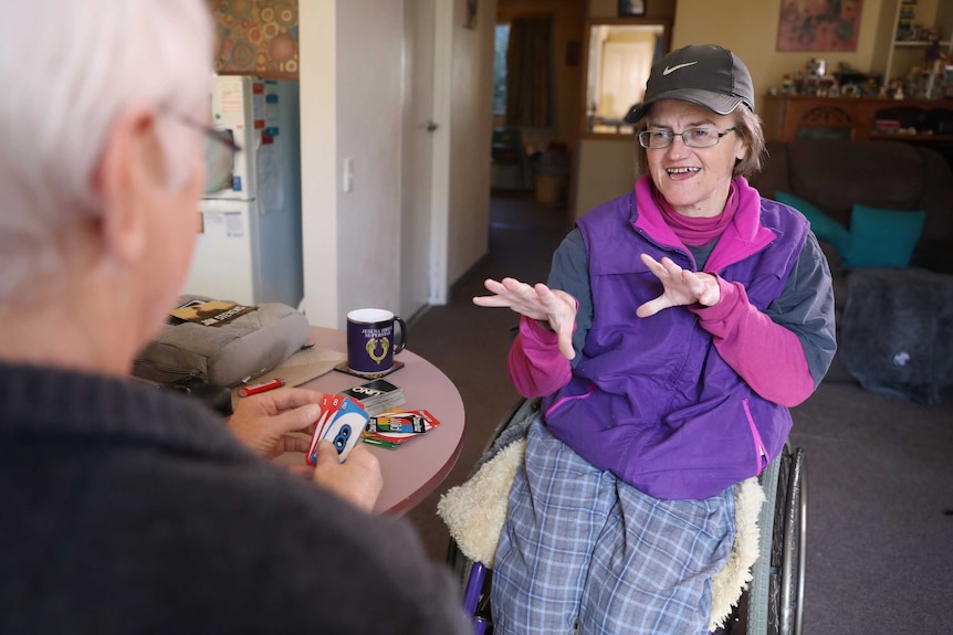 A middle-aged woman with hydrocephalus plays UNO with an older man around a table.