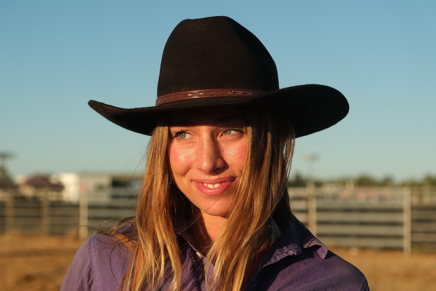 A young woman wearing a black hat looks into the distance, campdraft arena behind her