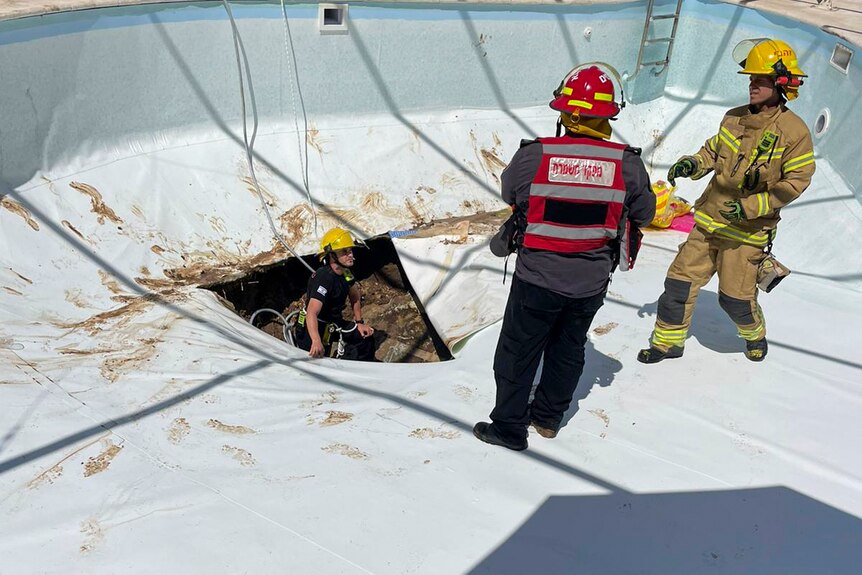 Two firemen are standing in the bottom of a pool, while another stands in a hole seemingly talking to each other.