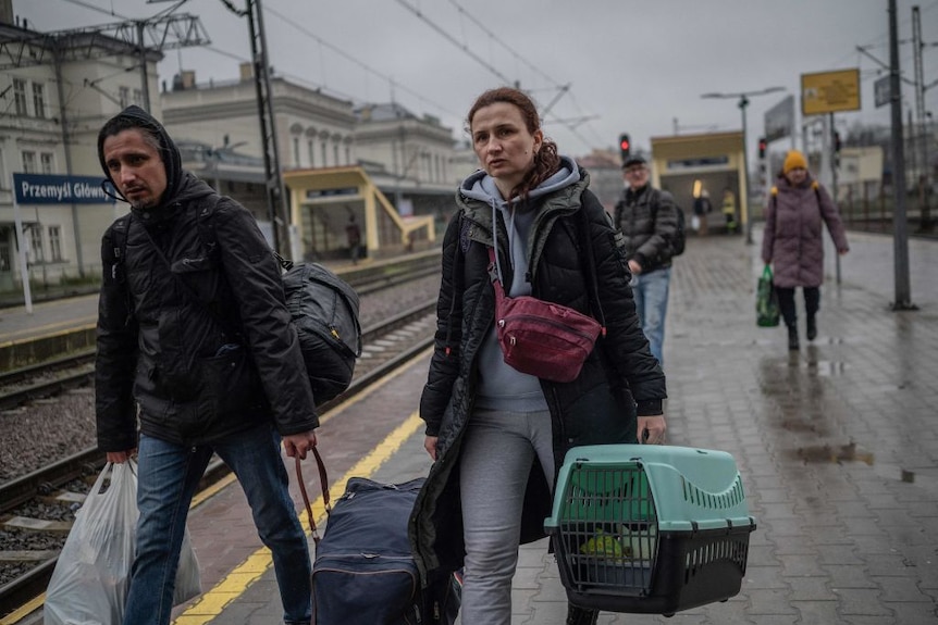 A middle-aged woman and man carry their luggage and cat along a train platform on an overcast and rainy day. 