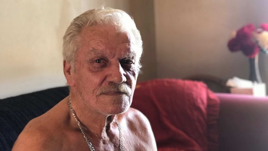 An elderly, moustachioed man wearing heavy gold chains around his neck sits shirtless on a couch.