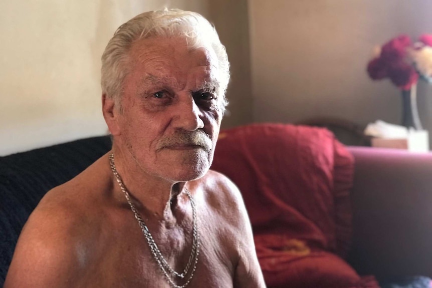 An elderly, moustachioed man wearing heavy gold chains around his neck sits shirtless on a couch.
