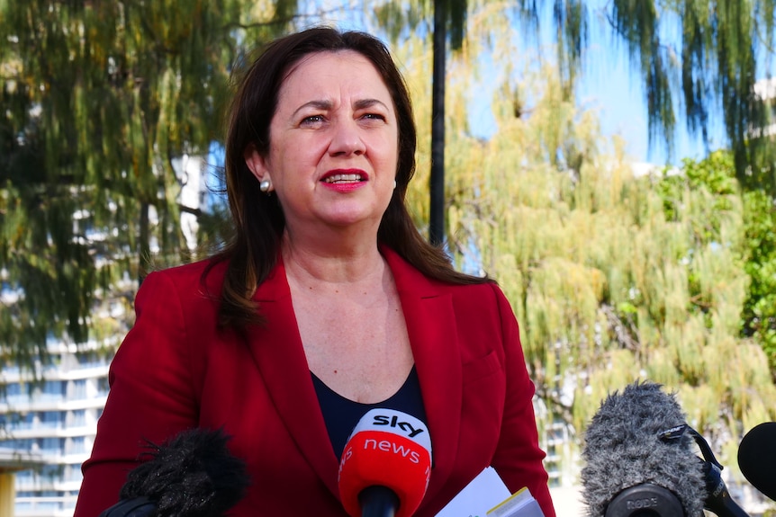 Premier in red jacket with black top, brown hair and earrings speaking in front of green trees and buildings