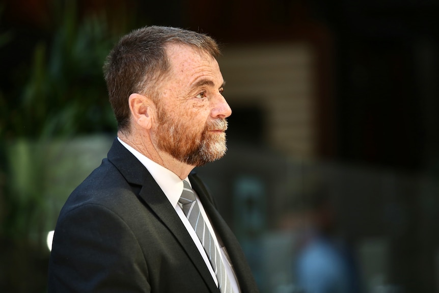 A man with an orange and grey beard but more brunette hair wearing a suit and tie walking towards right of frame.