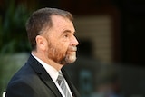 A man with an orange and grey beard but more brunette hair wearing a suit and tie walking towards right of frame.