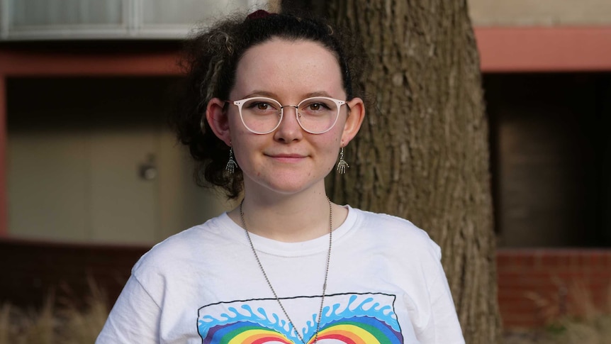 Findlay stares straight into the camera with a slight smile. She wears glasses and a white shirt with rainbow colours on it.