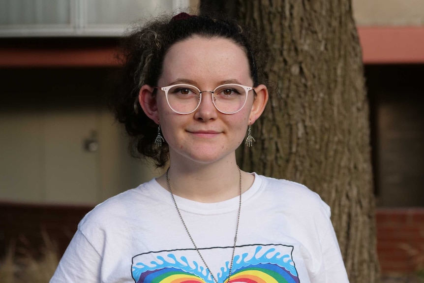 Findlay stares straight into the camera with a slight smile. She wears glasses and a white shirt with rainbow colours on it.
