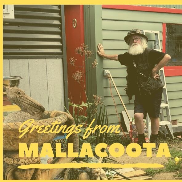 A man with white hair and beard leans against a timber house with Greetings from Mallacoota below