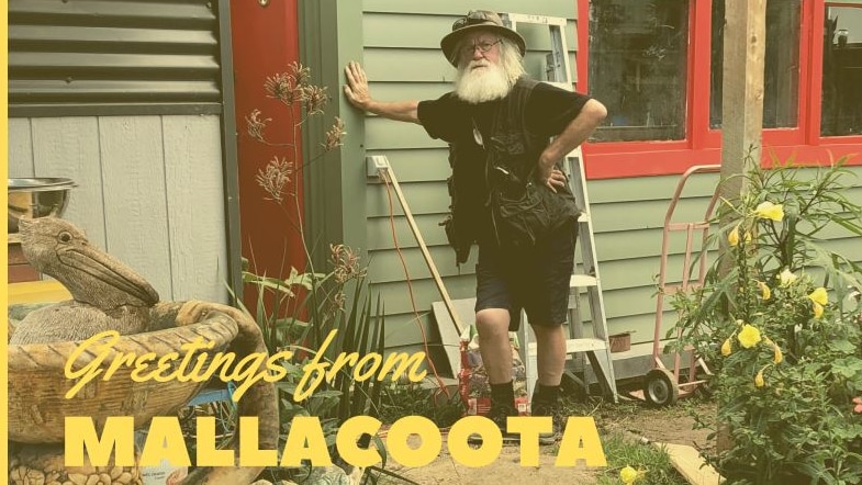 A man with white hair and beard leans against a timber house with Greetings from Mallacoota below