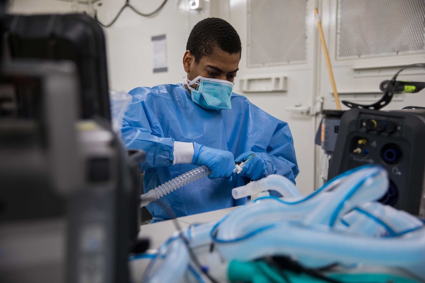 A man in blue scrubs with a mask on pulls on a plastic tube surrounded by equipment.