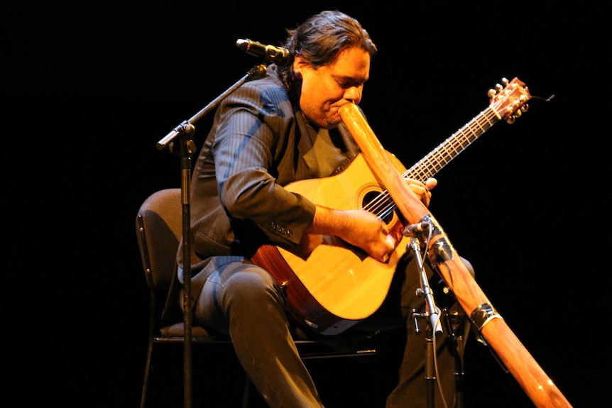 A dark haired man, formally attired, plays a didjeridu and a guitar at the same time.