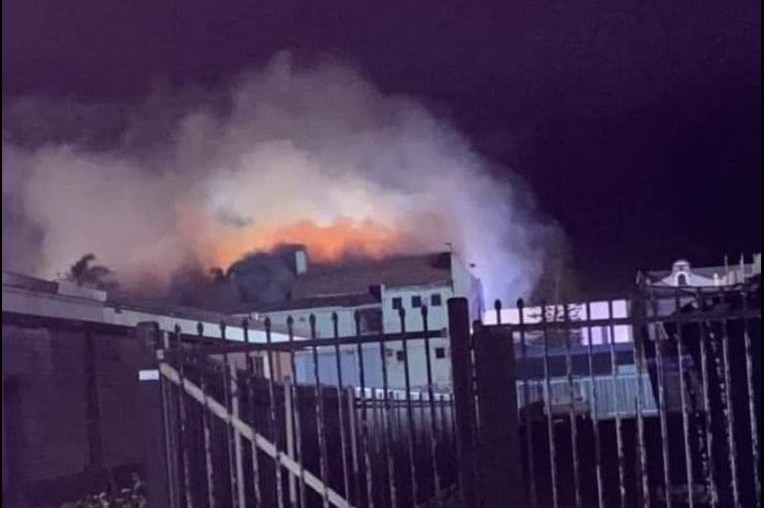 A photo, taken at night, shows the red glow of flames and smoke billowing from the roof of a building.