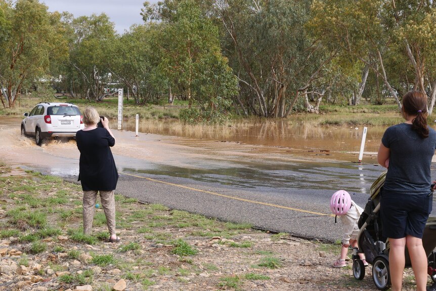 People stood on the side of the road watching as a car drives through brown water flowing over a road.
