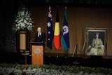 Anthony Albanese stands at a lectern with flags behind him and a painting of the Queen.