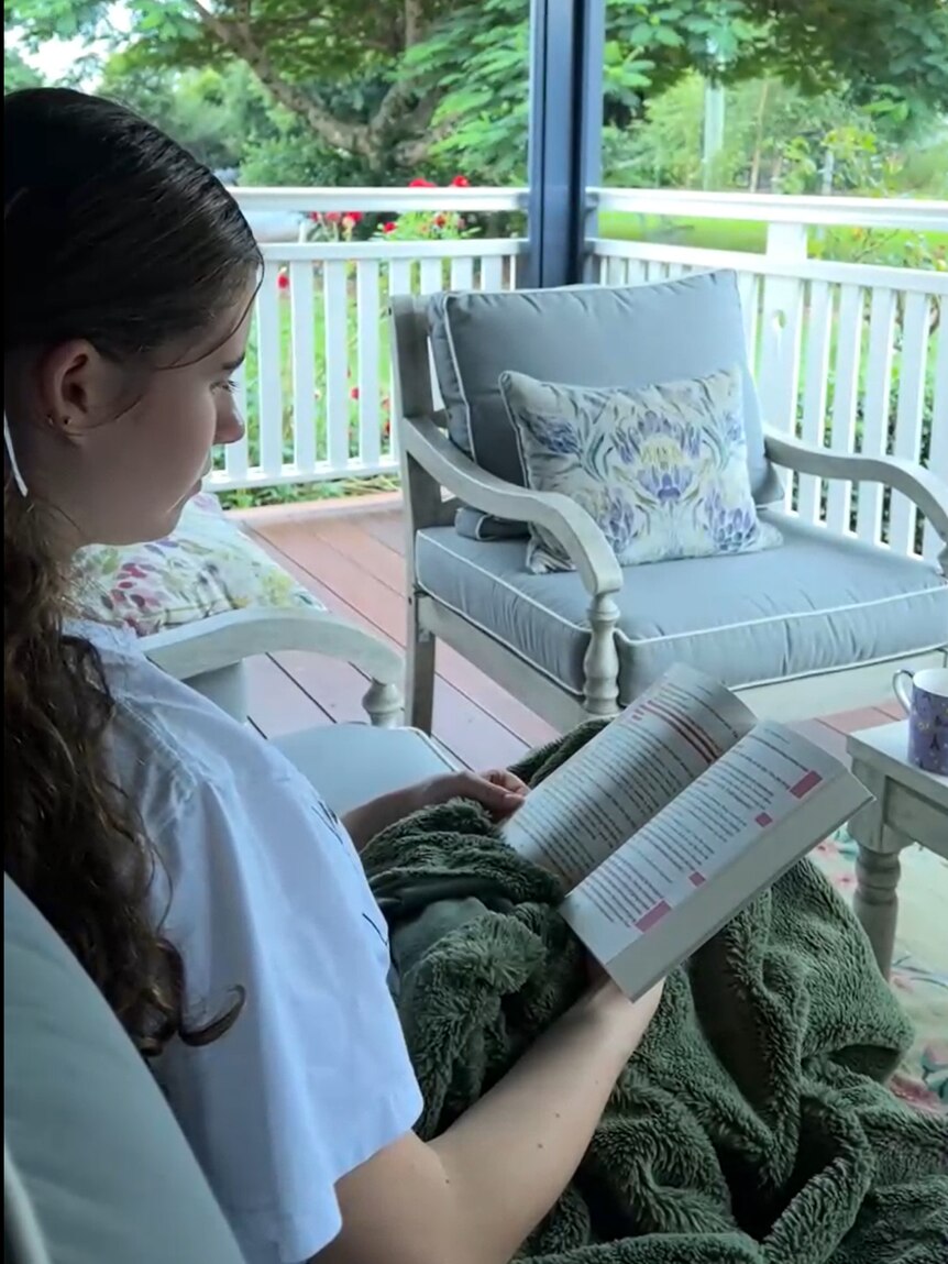 A young girl sits on outside on an armchair and reads a book.