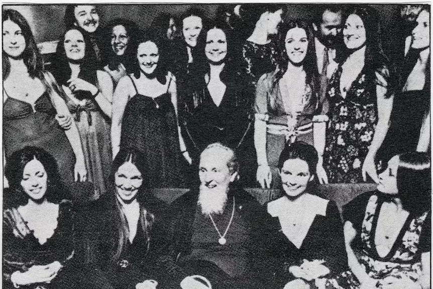 A black and white photo of an elderly bearded man posing for a photo with a large group of young women