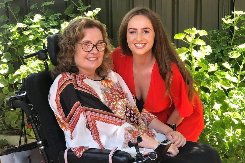 A woman in a wheelchair with a younger women crouched next to her, both smiling at the camera.