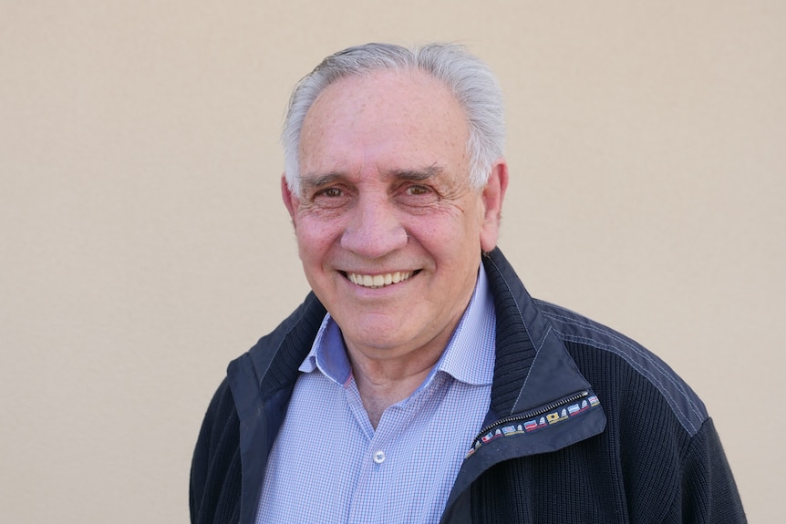 Paul Briggs wears a light blue shirt and dark coat, with grey hair and a big smile he looks into the camera.