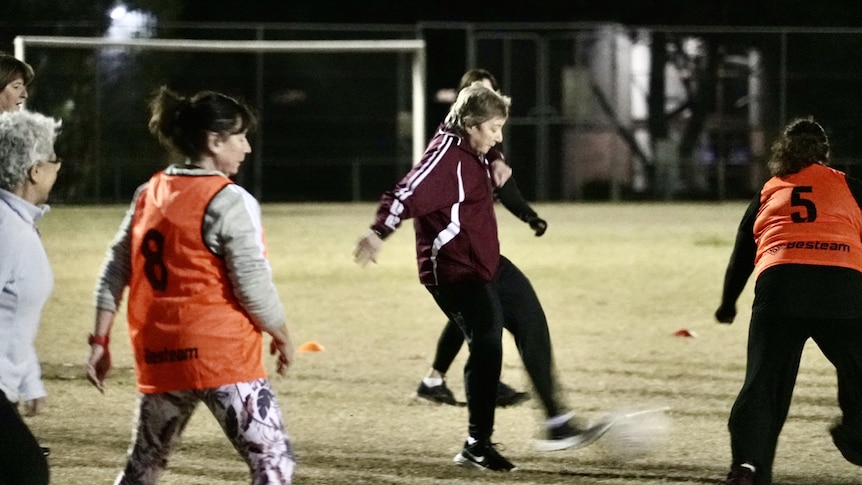 Women are seen playing football at a suburban pitch at night, light by flood lights