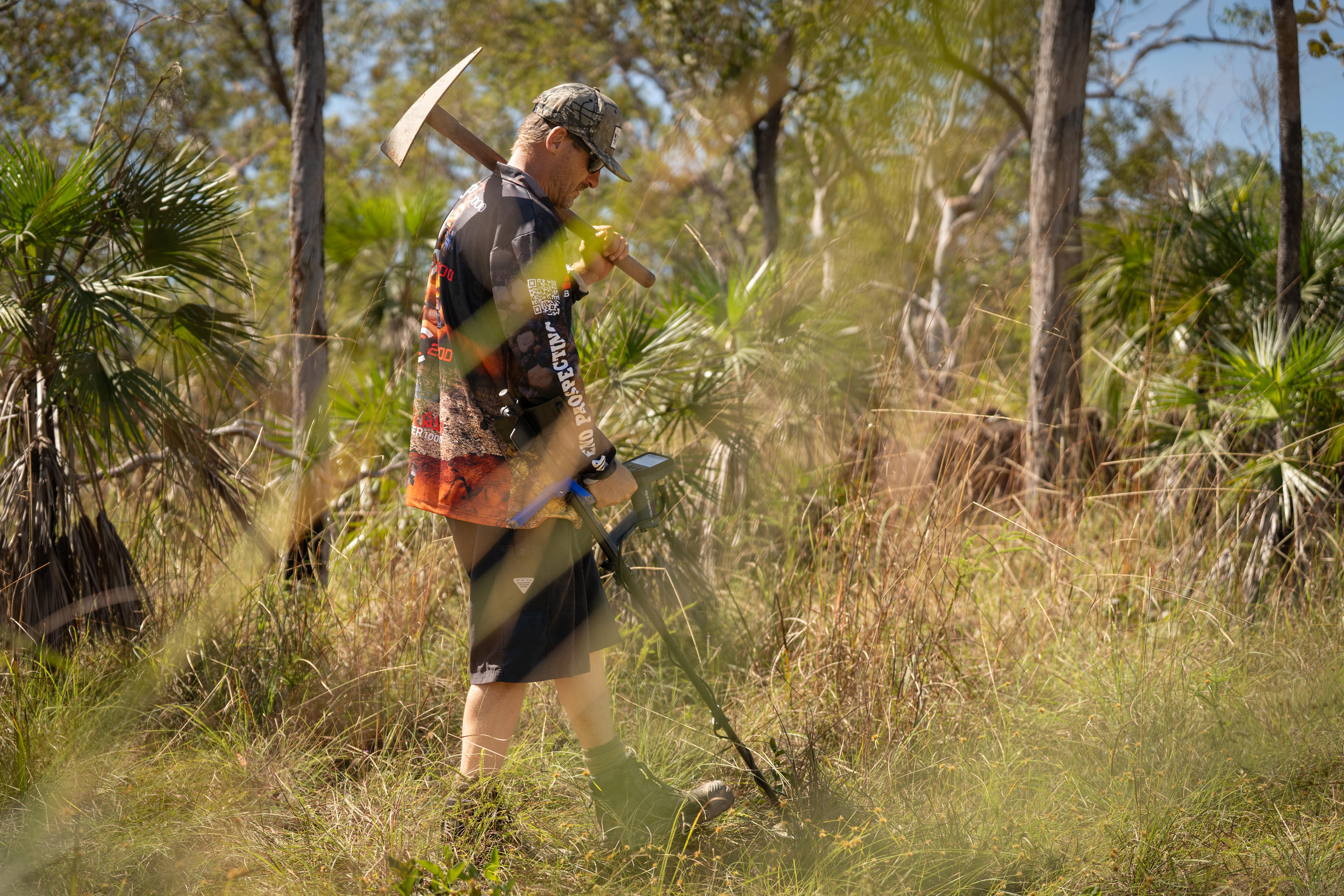 Man with mattock over shoulder and detector in the bush 