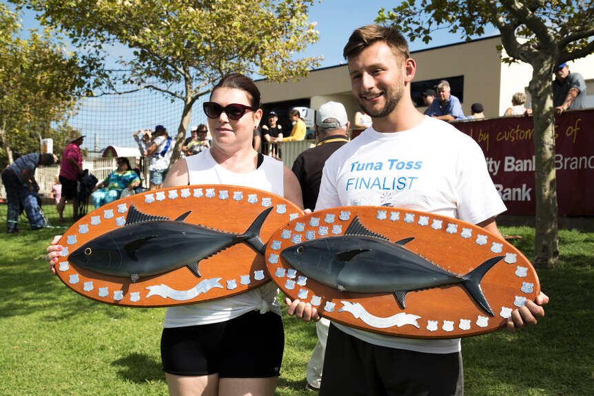 A brunette woman wearing sunglasses and a blonde man hold large plaques decorated with silver badges and replica tuna.