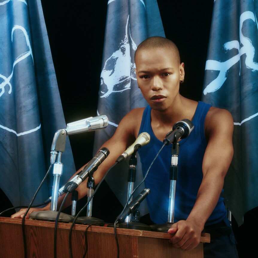 a person in blue tank top stands at a podium with microphones on it
