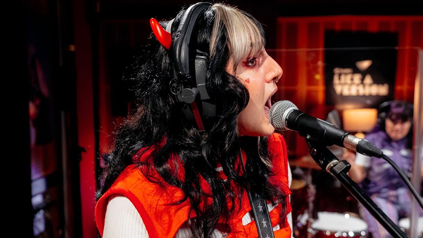 Person with a blonde fringe and black hair wearing devil horns and a red top, singing into a microphone