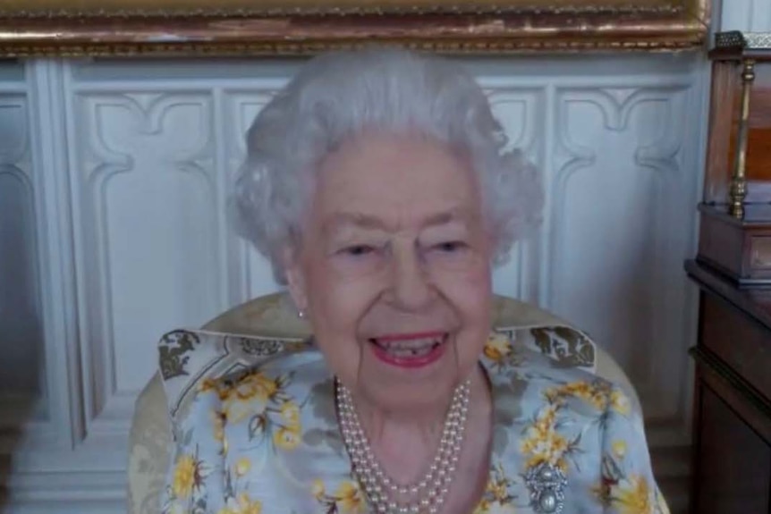 An elderly woman wearing a pearl necklace and a floral dress smiles