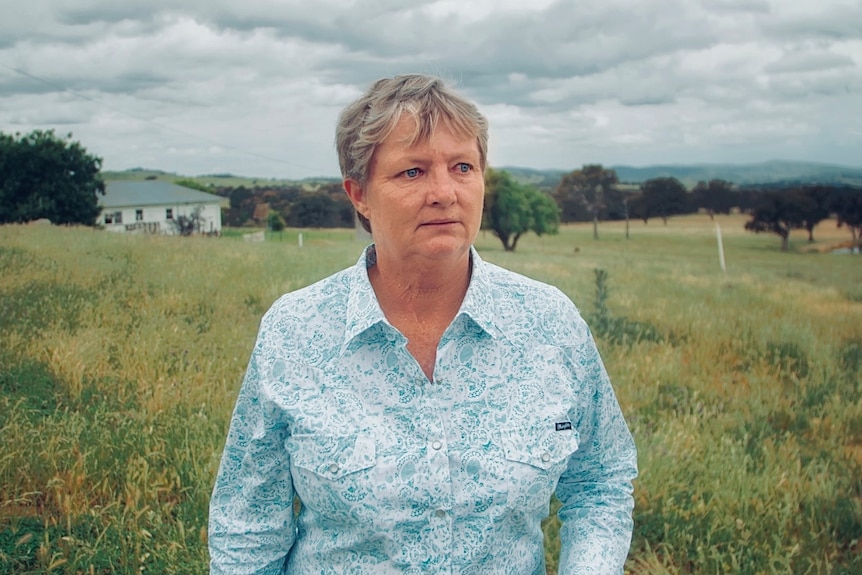 A middle-aged woman with short hair stands on a rural property and looks to the side of frame with a serious facial expression.