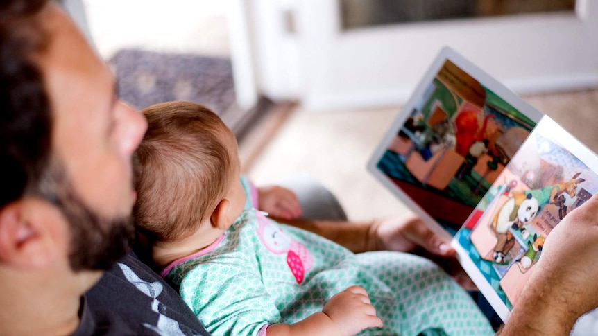 Father reads a childrens book to a young child in a chair.