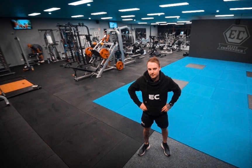 A man in his early 30s stands with his hands on his hips in a gym. There is fitness equipment in the background