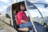 a man in a red work uniform sits in a helicopter on the ground, smiling at the camera