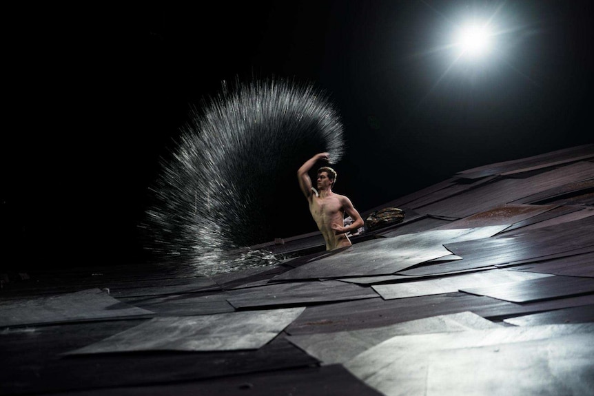 A naked man emerges in the middle of a stage splashing an arc of water.